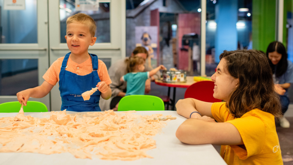 Wide shot of CMA Staff Member assisting with child in a smock and holding a paintbrush while playing with shaving cream on the table for Messy Thursdays.