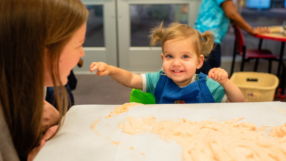 Close up of child smiling at their adult while playing in shaving cream for Messy Thursdays.